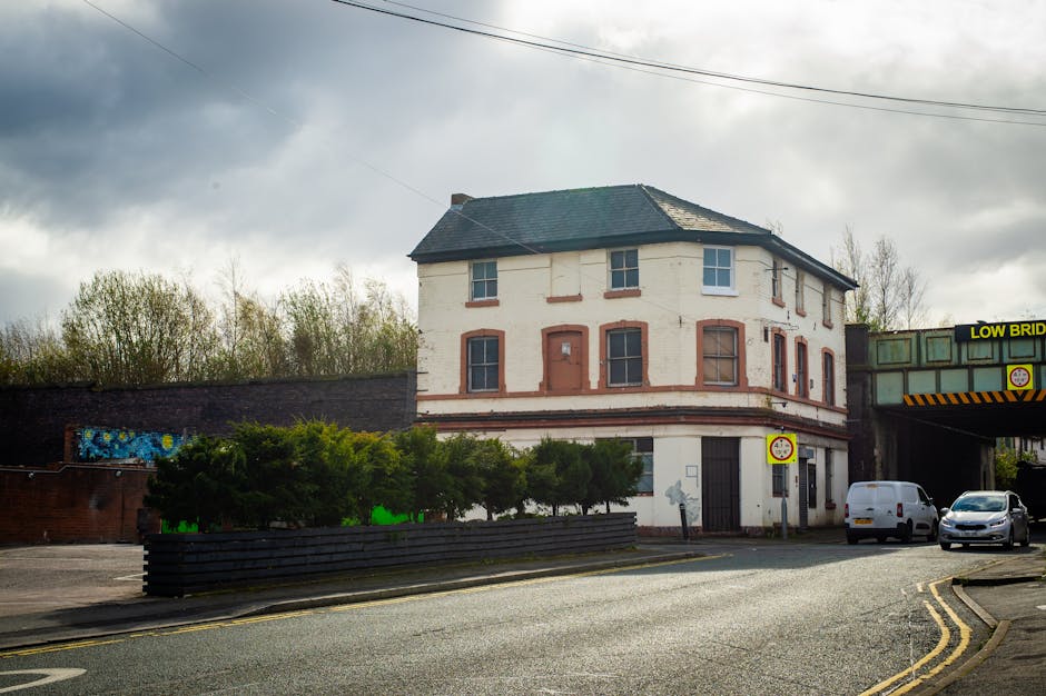 A three-storey, white-painted building with red brick window surrounds and a dark grey pitched roof, situated at a street corner near a bridge with a sign indicating a low bridge height. The building