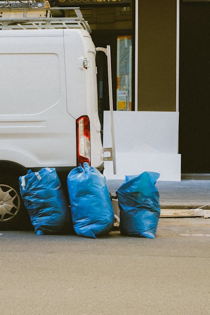 The rear section of a white van parked on a street curb, with three large blue moving bags made of durable plastic resting against its side, near the back wheel. The bags appear full and are used for 