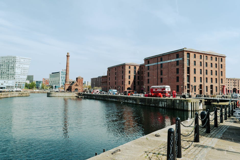 A wide view of the waterfront at Royal Albert Dock in Liverpool, showing a row of small boats and yachts moored along the stone-paved promenade, with black metal bollards and chains lining the edge. I