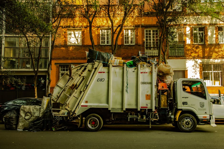 An extensive landfill site with a large, sweeping mound of mixed waste materials, including plastic, paper, and household rubbish, spread across the landscape. Several construction vehicles, including